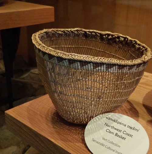 A woven clam basket from the Northwest Coast displayed on a wooden shelf with a descriptive label.
