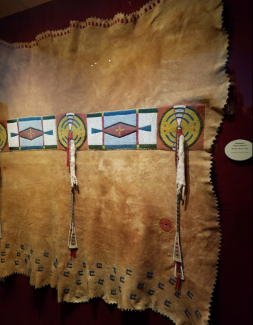 A decorative hide featuring colorful geometric patterns and tassels, displayed in a museum setting.