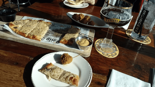 A wooden table with a plate of flatbread, a bowl of mustard, fried balls, and a glass of dark beer.