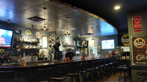 A cozy bar interior with a long counter, shelves of liquor, and a TV, featuring warm lighting and decorative signs.