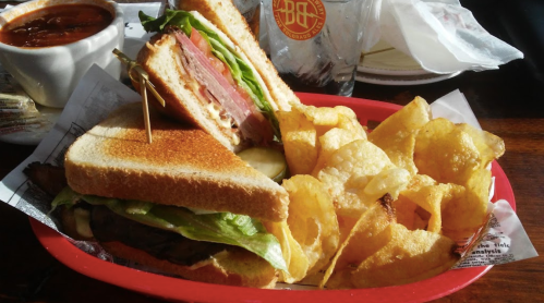 A toasted sandwich with layers of meat and lettuce, served with a side of crispy potato chips and a bowl of soup.