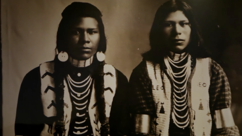 Two Indigenous individuals in traditional attire, adorned with jewelry and intricate patterns, posing for a portrait.