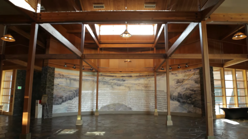Interior of a spacious wooden building with a mural and large windows, featuring natural light and a stone floor.