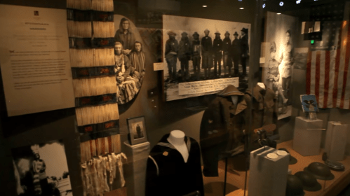 Display of historical artifacts, including military uniforms, photographs, and Native American items in a museum setting.