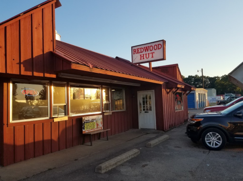 Exterior of Redwood Hut restaurant with a red wooden facade, open sign, and parked cars in the lot during sunset.
