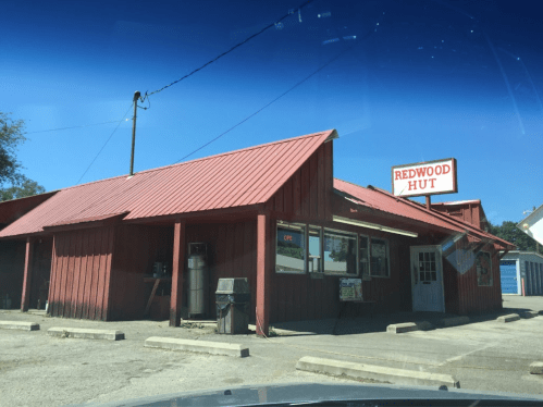 A red wooden building with a metal roof, featuring a sign that reads "Redwood Hut" and a small outdoor area.