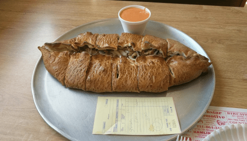 A large calzone on a metal plate, filled with ingredients, accompanied by a small cup of sauce and a receipt.