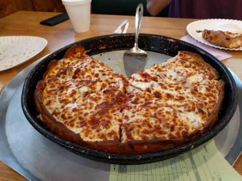 A deep-dish pizza with a golden crust and melted cheese, partially sliced, on a metal tray with a serving spatula.
