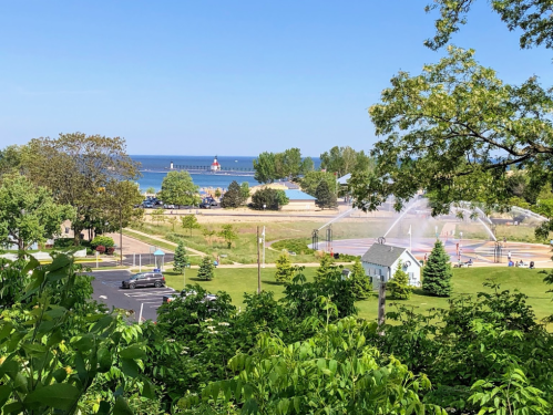 A scenic view of a beach with a lighthouse, fountains, and green trees under a clear blue sky.