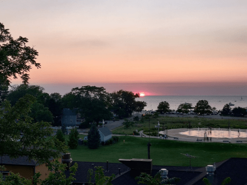 A serene sunset over a lake, with trees and a park in the foreground, casting a warm glow in the sky.