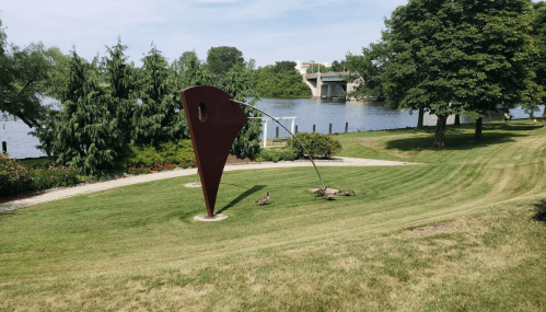 A park scene featuring a large metal sculpture, green grass, and geese near a river with trees in the background.