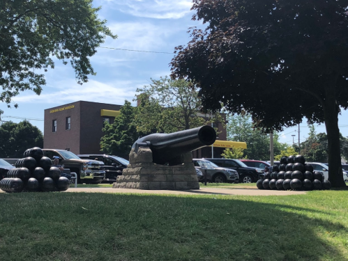 A historic cannon on a stone base, surrounded by cannonballs, with a building and trees in the background.