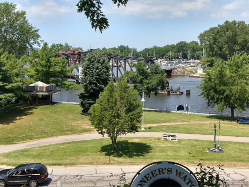 A scenic view of a river with a bridge, boats, and green trees under a blue sky.