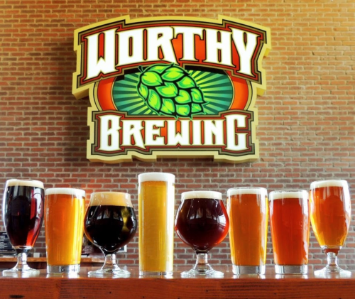 A variety of beer glasses in front of a "Worthy Brewing" sign on a brick wall.