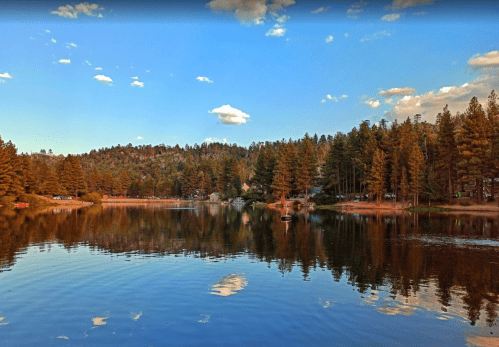 A serene lake surrounded by tall pine trees and a clear blue sky with fluffy clouds reflecting on the water's surface.