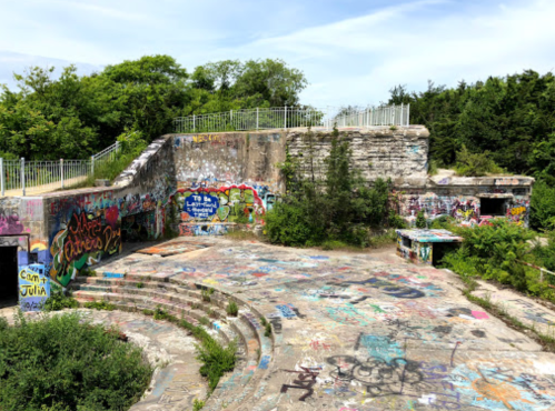 Abandoned outdoor space with overgrown vegetation and colorful graffiti on walls and ground under a blue sky.