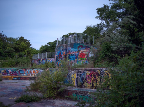 Abandoned structure covered in colorful graffiti, surrounded by overgrown vegetation and trees under a twilight sky.