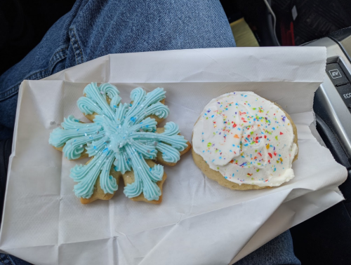 Two decorated cookies on a white paper napkin: one shaped like a snowflake with blue icing, the other a round cookie with white frosting and sprinkles.
