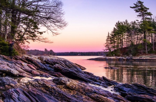 A serene lakeside scene at sunset, featuring rocky shores and trees silhouetted against a colorful sky.