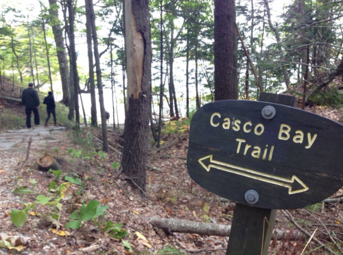 Two people stand near a sign for the Casco Bay Trail, surrounded by trees and a glimpse of water in the background.