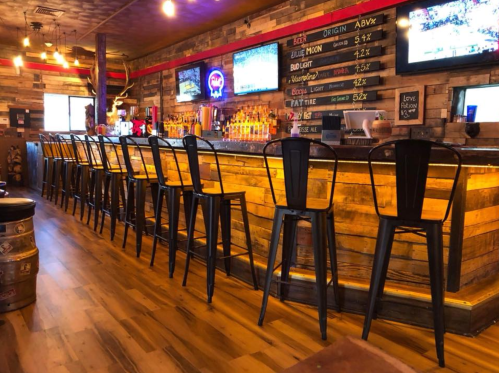 A rustic bar with wooden decor, tall black chairs, and multiple beer taps, illuminated by warm lighting.