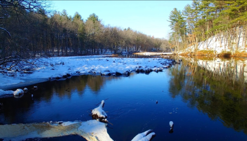 A serene winter landscape featuring a calm river surrounded by snow-covered banks and trees under a clear blue sky.