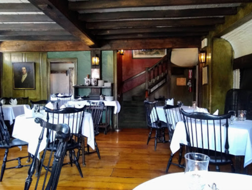 Cozy restaurant interior with wooden beams, tables set with white linens, and a staircase in the background.