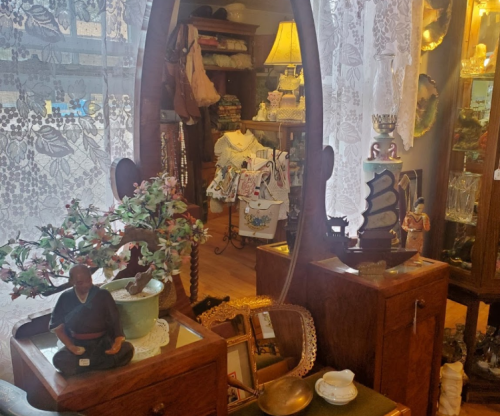 A vintage shop interior featuring a large mirror, decorative items, and various antiques displayed on wooden furniture.