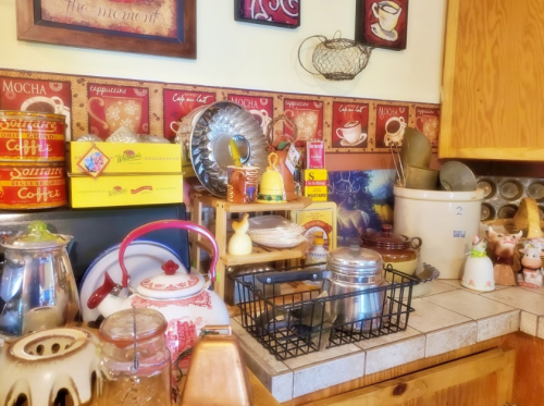 A cozy kitchen counter filled with vintage coffee pots, jars, and decorative items against a warm wooden backdrop.