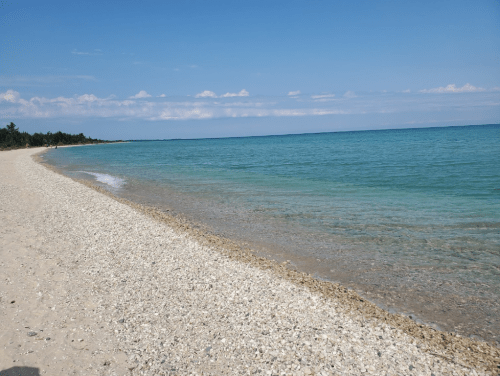 A serene beach with a sandy shore and clear blue water under a bright sky, dotted with a few clouds.