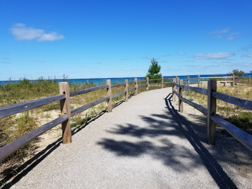A sandy path lined with wooden fences leads to a blue lake under a clear sky with a few clouds.