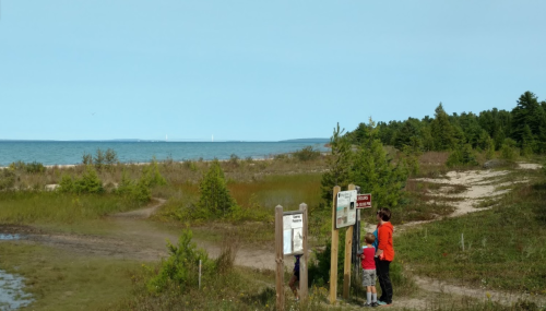 A child and an adult stand by a trail sign near a beach, with water and trees in the background under a clear sky.