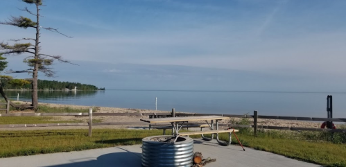 Serene beach view with calm water, a picnic table, and a fire pit surrounded by trees and a wooden fence.