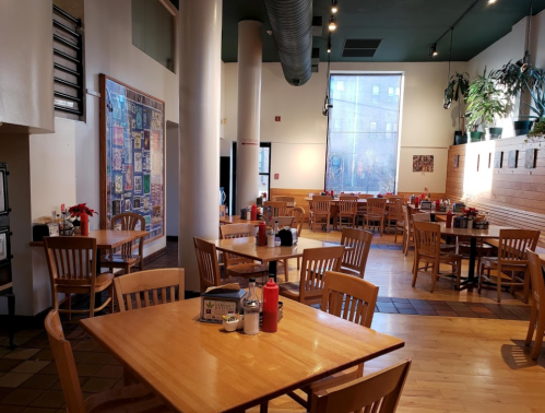 A spacious, empty restaurant with wooden tables, chairs, and a large window, featuring plants and decor on the walls.