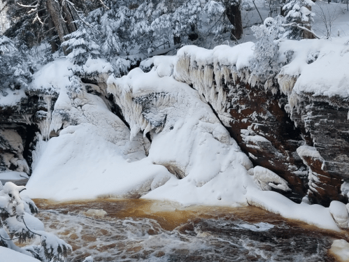 Snow-covered cliffs with icicles above a flowing river, surrounded by trees in a winter landscape.