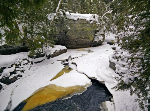 A snowy landscape featuring a river with ice and snow-covered rocks, surrounded by evergreen trees.