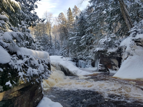 A snowy landscape featuring a waterfall surrounded by trees and rocky cliffs, with frothy water flowing downstream.