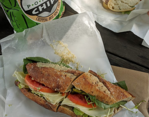 A sandwich with lettuce, tomato, and sprouts on a wooden table, next to a bag of chips.