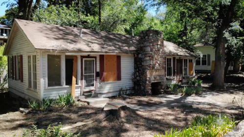 A rustic house with boarded windows and a stone chimney, surrounded by trees and overgrown landscaping.