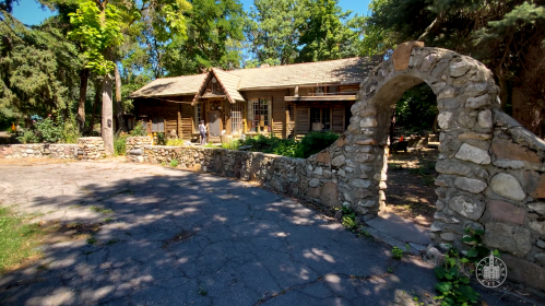 A rustic log cabin surrounded by trees, with a stone archway and a paved path leading to the entrance.