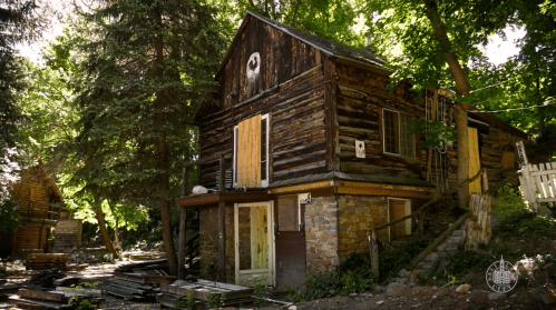 A rustic log cabin surrounded by trees, featuring a stone foundation and wooden porch, in a natural setting.