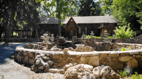 A rustic wooden cabin surrounded by greenery and a stone fountain in a serene outdoor setting.