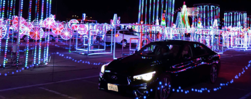 A black car drives through a colorful display of holiday lights in a festive outdoor setting.