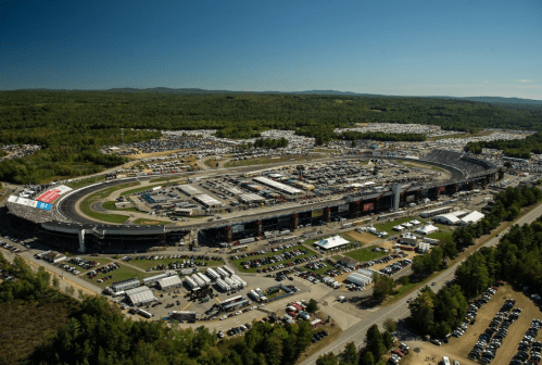 Aerial view of a busy racetrack surrounded by trees and parking lots, with spectators and vehicles visible.