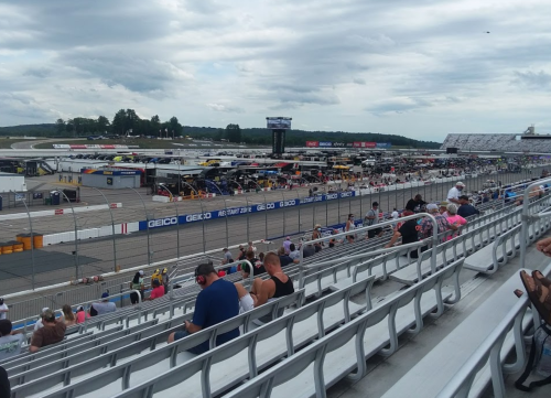 A view of a racetrack with empty grandstands and parked race cars in the background under a cloudy sky.