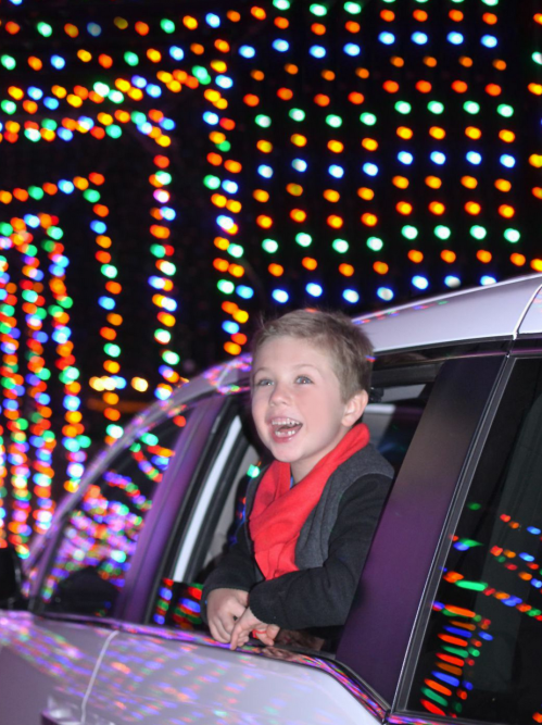 A joyful child leans out of a car window, surrounded by colorful holiday lights.