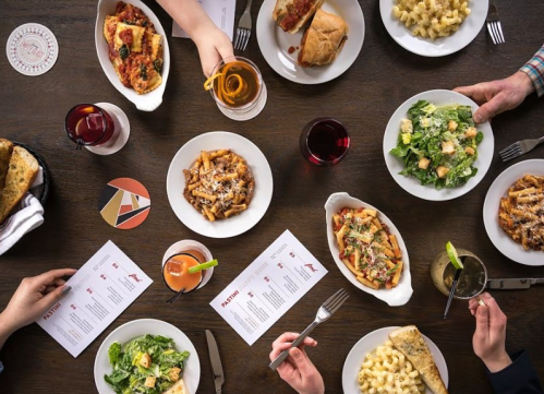 A table spread with various pasta dishes, salads, drinks, and menus, with hands reaching for the food.