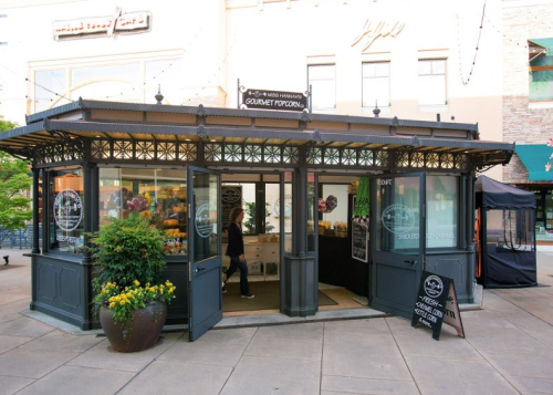 A charming kiosk selling gourmet popcorn, featuring large windows and a welcoming entrance, surrounded by greenery.
