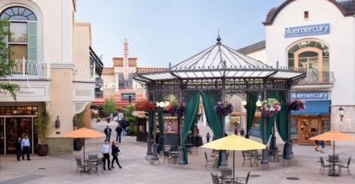 A vibrant outdoor shopping area with a gazebo, colorful flowers, and people enjoying the space under sunny skies.
