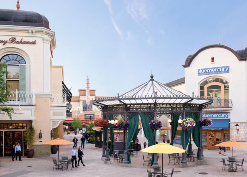 A vibrant outdoor shopping area with a gazebo, colorful flowers, and people enjoying the space under sunny skies.